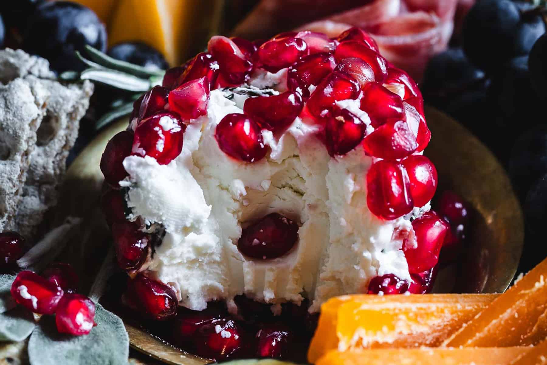 A cheese ball topped with pomegranate seeds, surrounded by crackers and cheese slices.