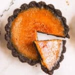 Mini Pumpkin Brûlée Tarts with a dark chocolate crust, one slice cut and slightly pulled out on a white surface.