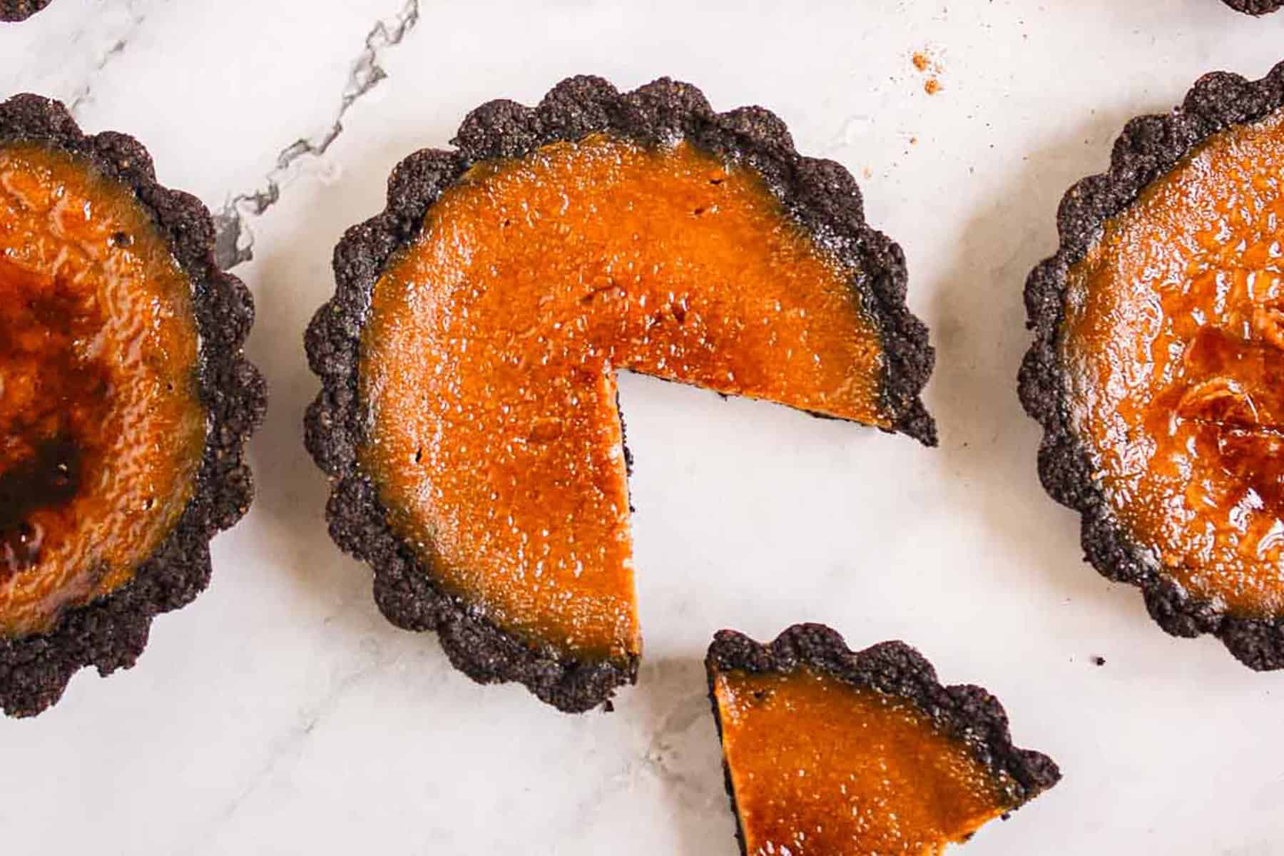 Three Pumpkin Br&ucirc;l&eacute;e Tarts with caramelized tops and dark crusts rest on a white surface, one tart with a slice cut out.