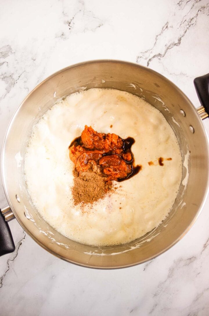 A metal pot with frothy liquid, tomato paste, spices, and dark sauce sits on a marble countertop, next to a platter of Pumpkin Rice Krispies Donuts.