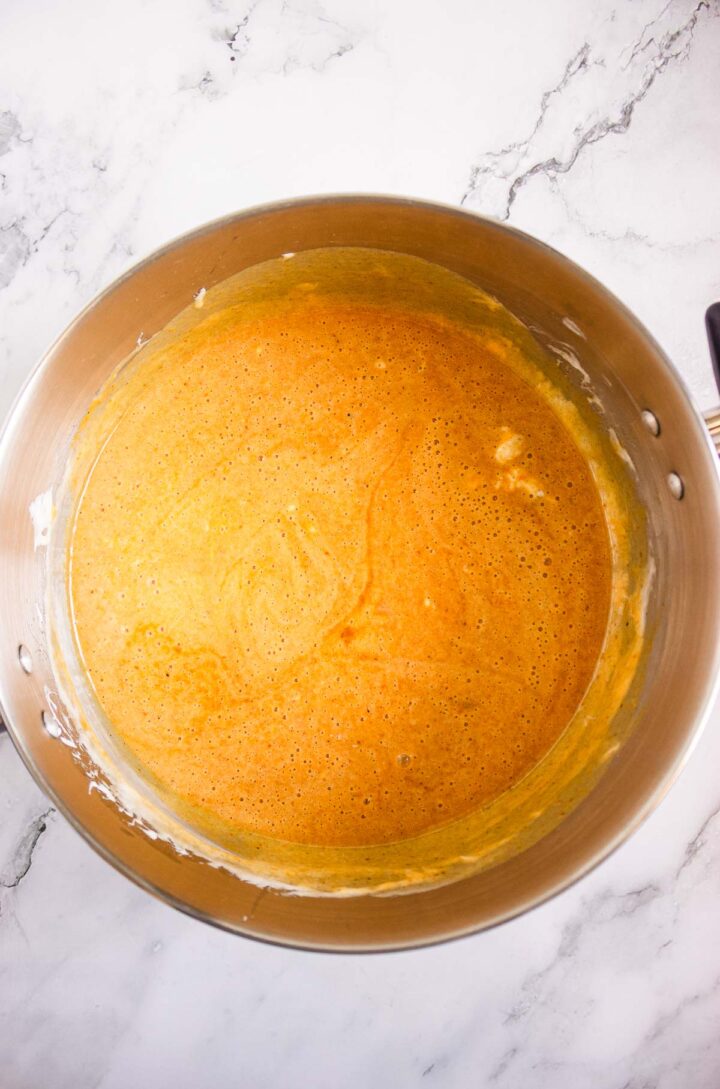 Stainless steel bowl filled with orange pumpkin pie batter for Pumpkin Rice Krispies Donuts on a white marble countertop.