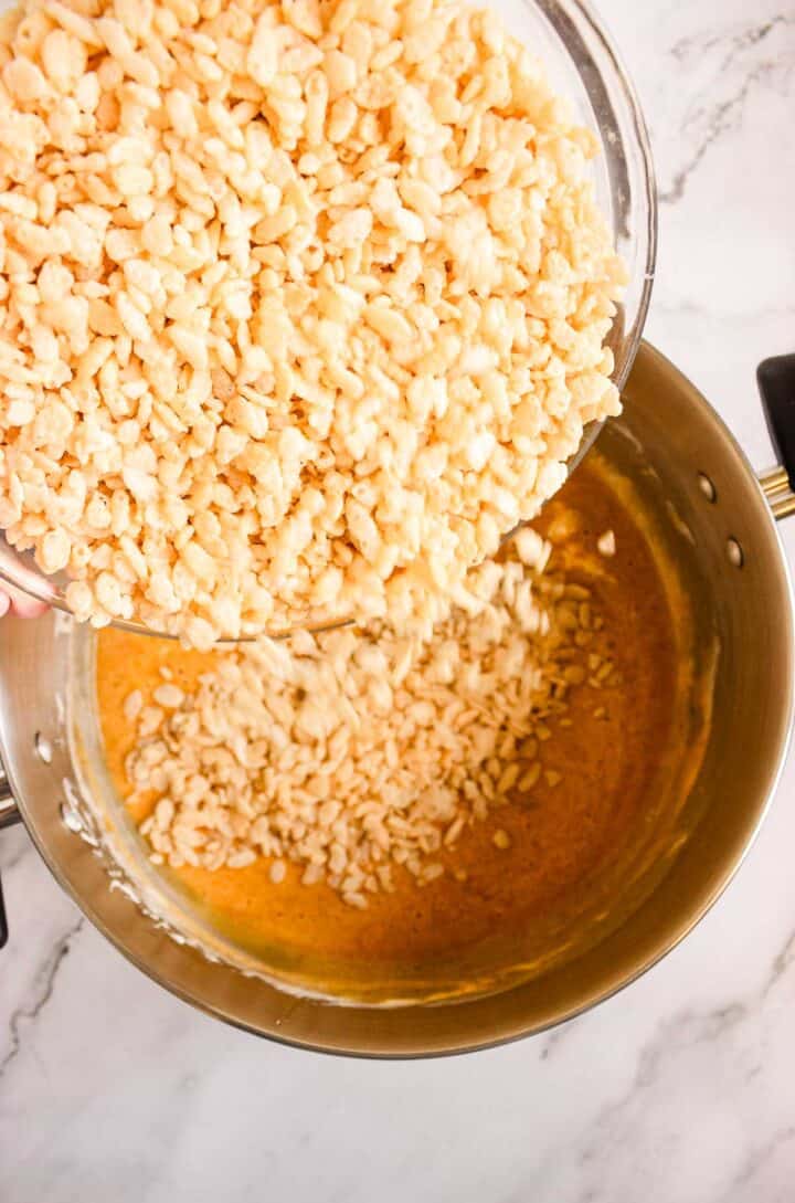 A bowl of rice cereal, perfect for making Pumpkin Rice Krispies Donuts, is being poured into a pot of melted marshmallow mixture on a marble countertop.