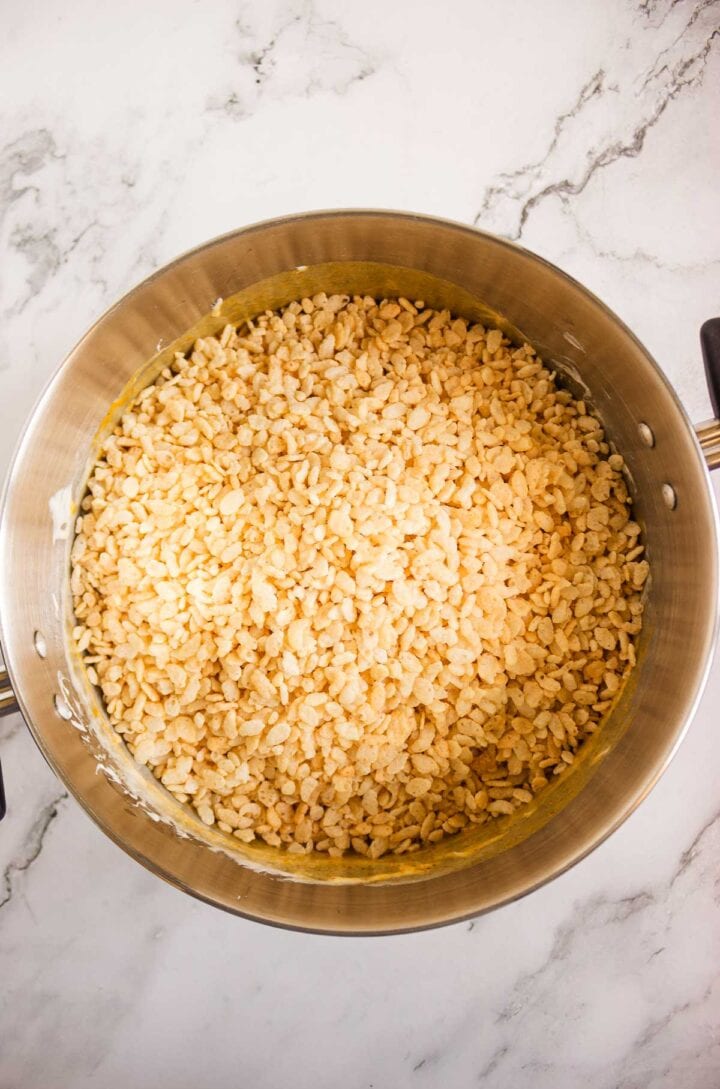 A pot filled with Rice Krispies cereal and melted marshmallows, perfect for making festive Pumpkin Rice Krispies Donuts, sits on a marble countertop.