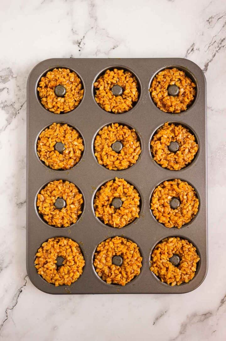 A donut pan filled with twelve round, golden Pumpkin Rice Krispies Donuts sits on a marble countertop.
