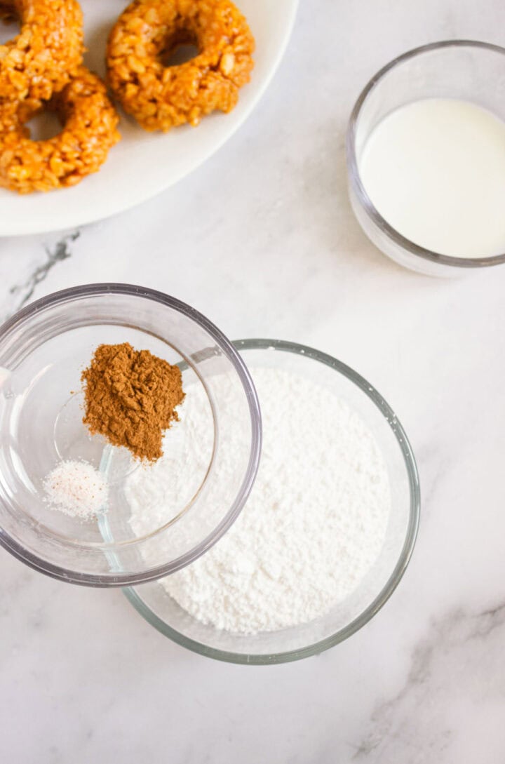 Three bowls with flour, cinnamon, and milk on a marble counter; plate of Pumpkin Rice Krispies Donuts in background, top left.