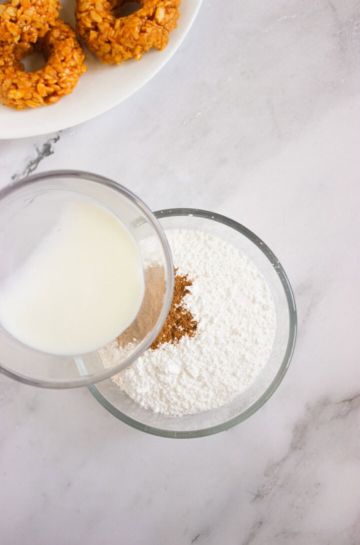 A bowl of flour and spices with milk being poured in, next to a plate of Pumpkin Rice Krispies Donuts on a marble surface.