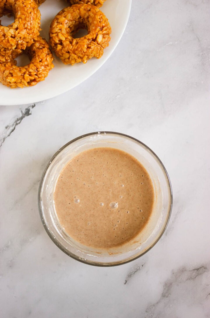 A bowl of light brown glaze sits next to a plate of Pumpkin Rice Krispies Donuts, all arranged on a marble surface.