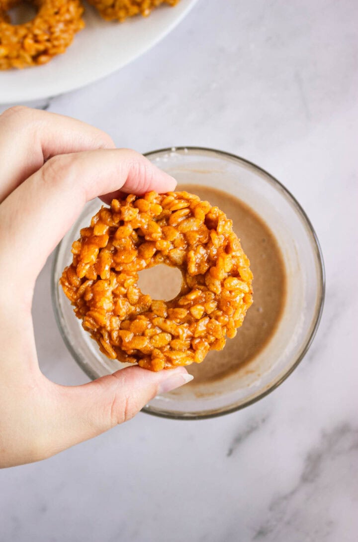 A hand holds a round Pumpkin Rice Krispies Donut above a bowl of light brown dipping sauce on a marble surface.