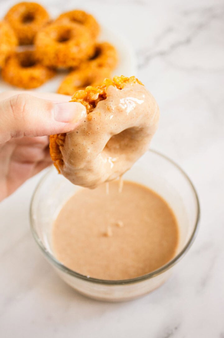 A hand dips a Pumpkin Rice Krispies Donut into a bowl of glaze, with more donuts on a plate in the background.