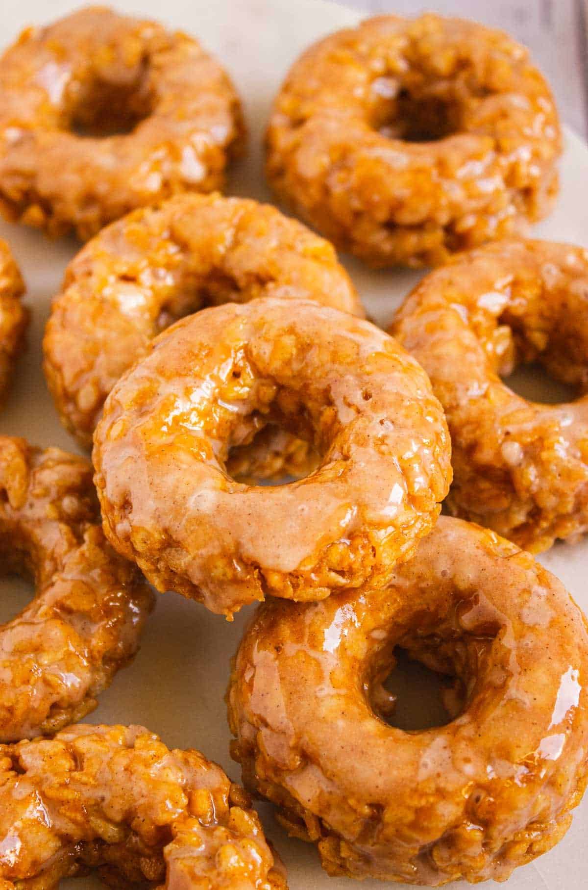 A pile of glazed apple fritters stacked closely together on a light surface, beside some Pumpkin Rice Krispies donuts.