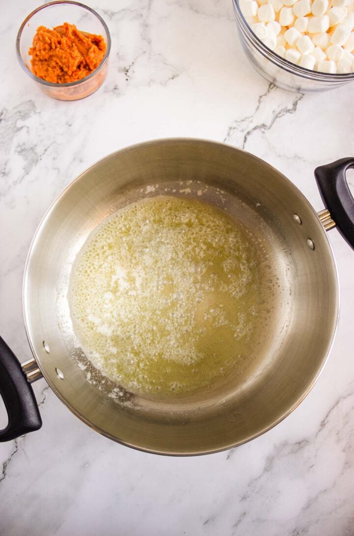 Melted butter in a saucepan, with bowls of marshmallows and orange mixture for Pumpkin Rice Krispies Donuts on a marble countertop.