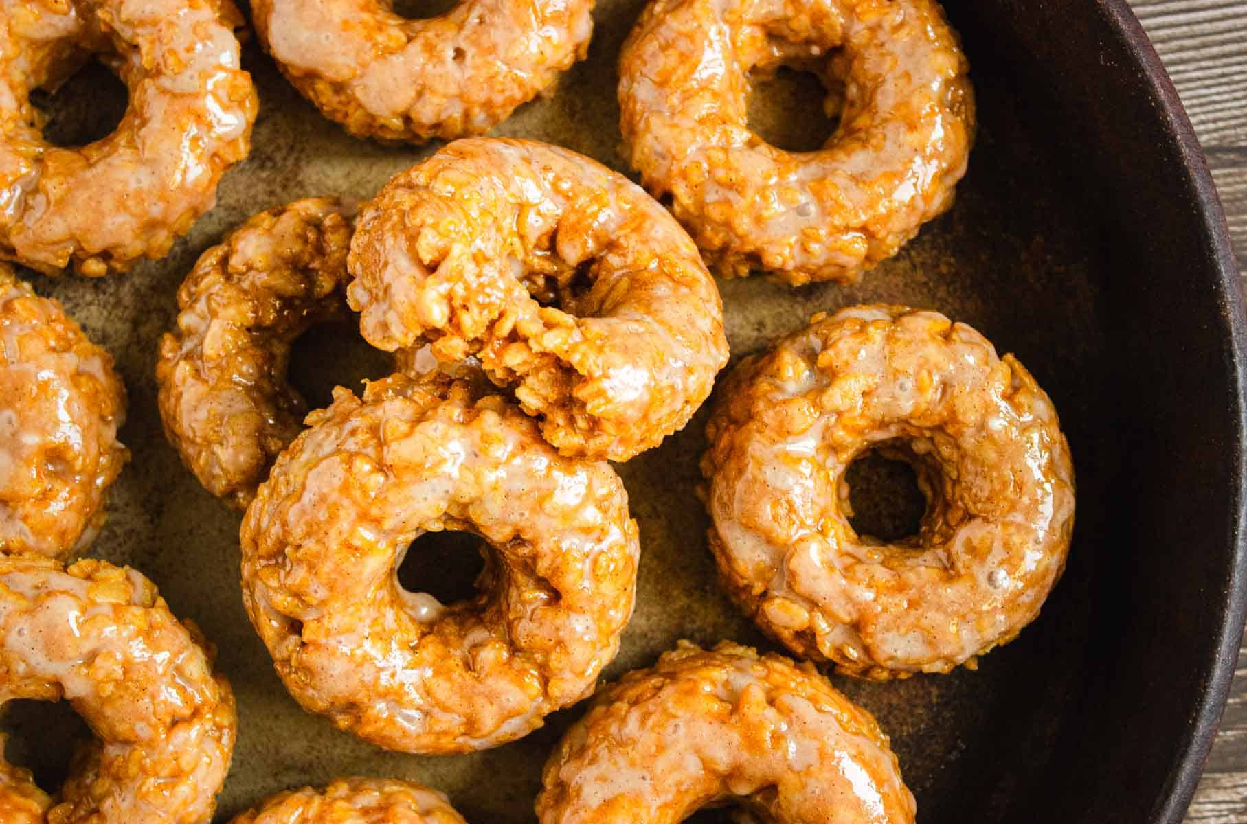 Close-up of glazed Pumpkin Rice Krispies Donuts arranged in a pan, with one donut partially broken.