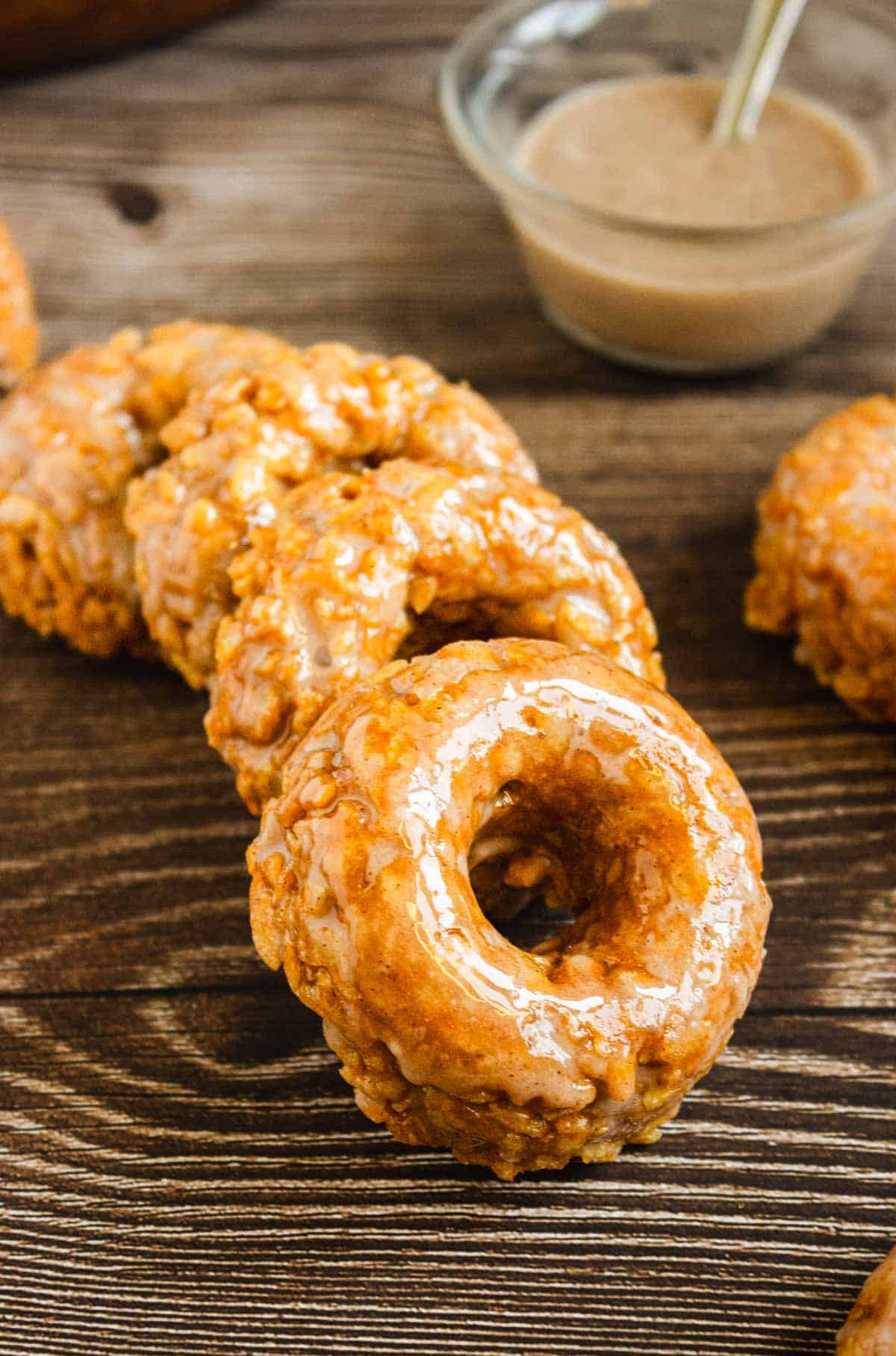 Glazed apple fritters stacked on a wooden surface with a bowl of brown dipping sauce in the background, alongside Pumpkin Rice Krispies donuts for a delightful autumn treat.