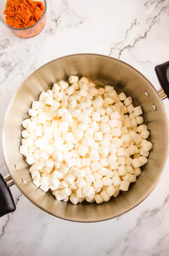 Large pot filled with mini marshmallows, alongside a bowl of mashed sweet potatoes and Pumpkin Rice Krispies Donuts on a marble counter.