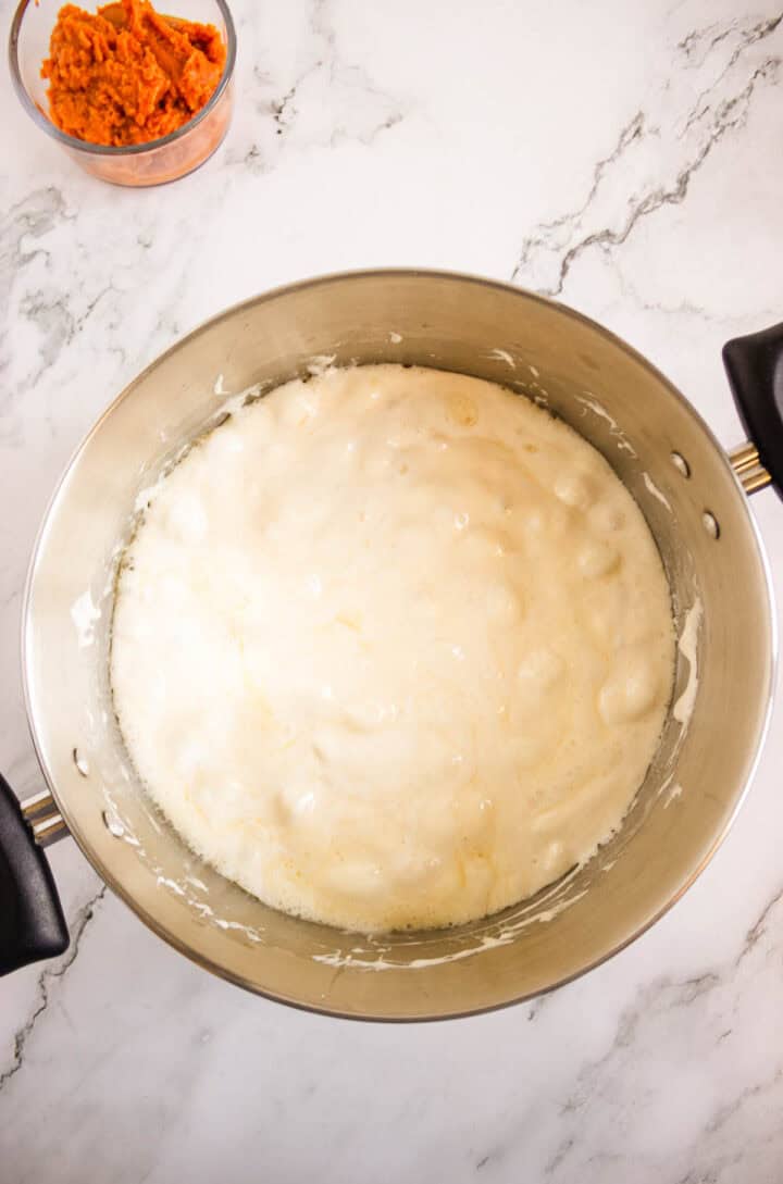 A metal mixing bowl of bubbly yeast mixture sits next to a small bowl of mashed sweet potatoes on a marble counter, ready for baking Pumpkin Donuts.