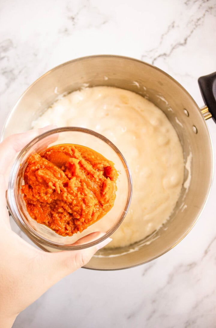 Hand holding a bowl of pumpkin puree above a mixing bowl with batter on a marble surface, preparing for Pumpkin Rice Krispies Donuts.