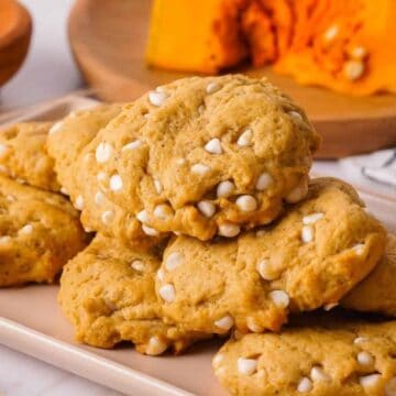 A plate of pumpkin cookies with white chocolate chips, next to a glass of milk and sliced pumpkin in the background.