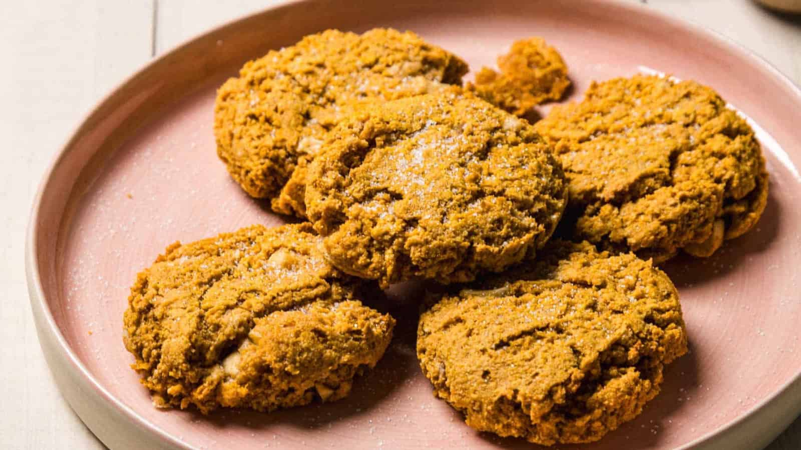 Four pumpkin cookies stacked on a pink plate, surrounded by pinecones and a baking tray in the background.