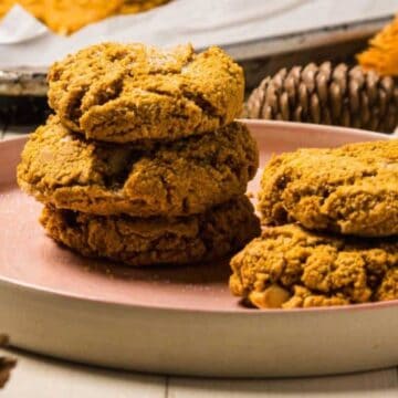 Four pumpkin cookies stacked on a pink plate, surrounded by pinecones and a baking tray in the background.