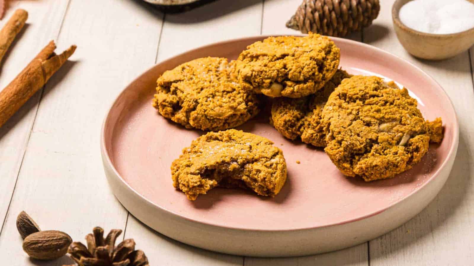 Four pumpkin cookies stacked on a pink plate, surrounded by pinecones and a baking tray in the background.