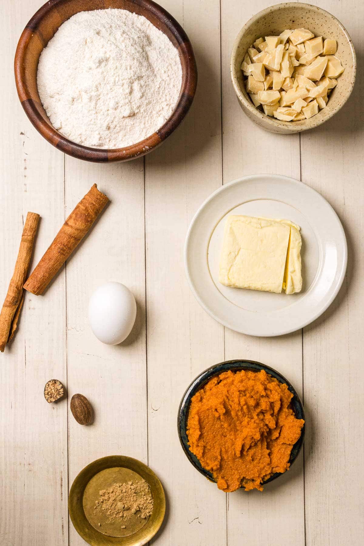 Baking ingredients on a wooden table for Pumpkin and White Chocolate Chip Cookies: flour, white chocolate, butter, pumpkin puree, egg, spices, and cinnamon sticks.
