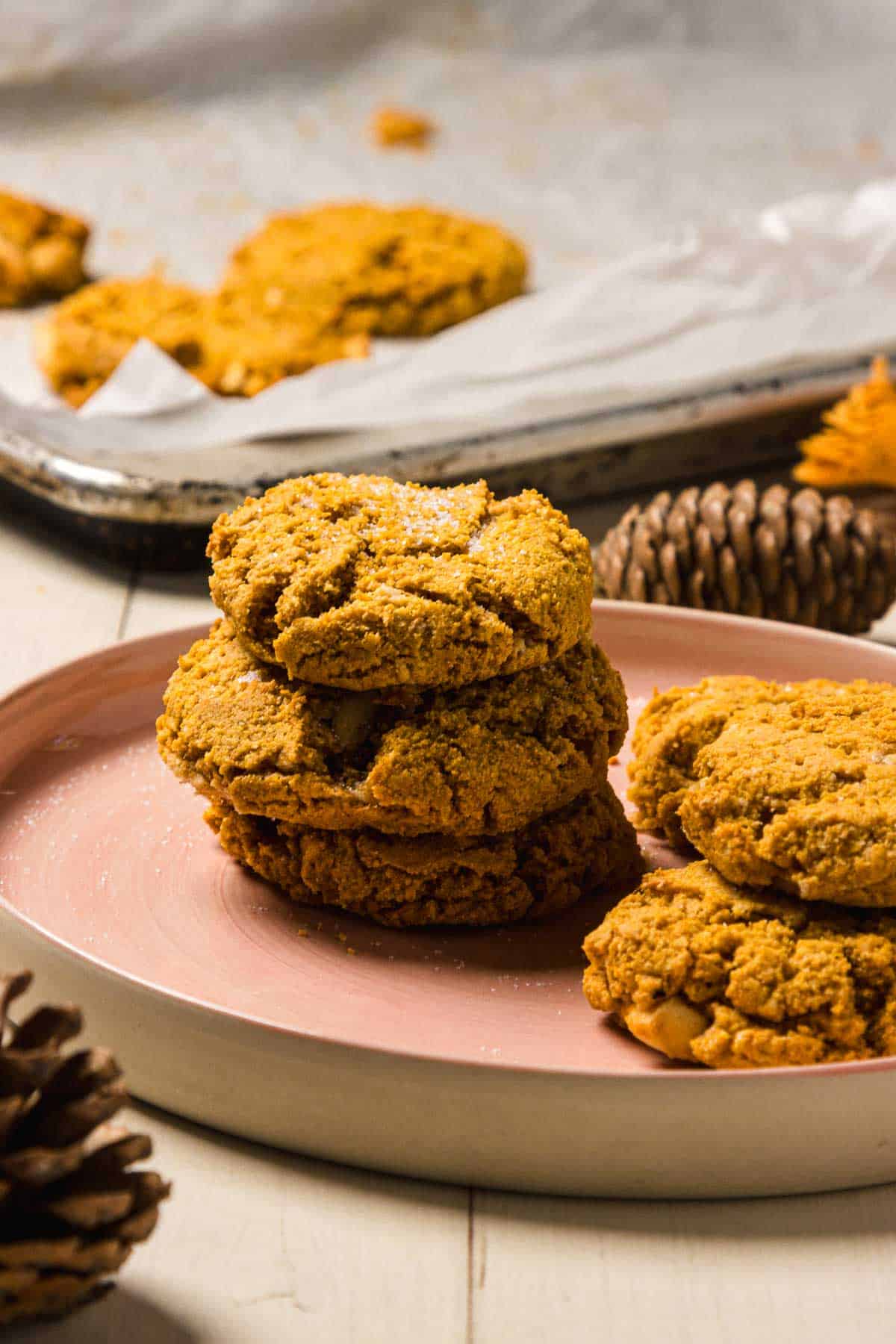 A stack of Pumpkin and White Chocolate Chip Cookies on a pink plate, with more cookies and pinecones in the background.