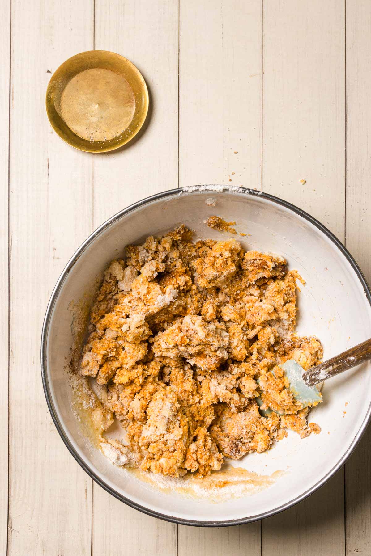 A bowl of crumbly Pumpkin and White Chocolate Chip Cookies dough being mixed with a spatula on a light wooden surface; empty dish nearby.