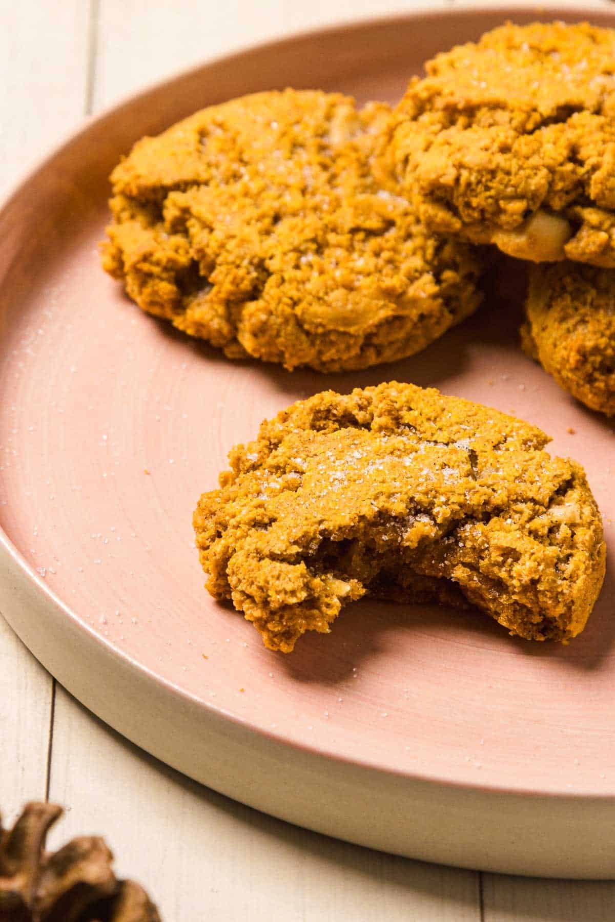 A plate of three Pumpkin and White Chocolate Chip Cookies, one with a bite taken out, sits on a light wooden table.