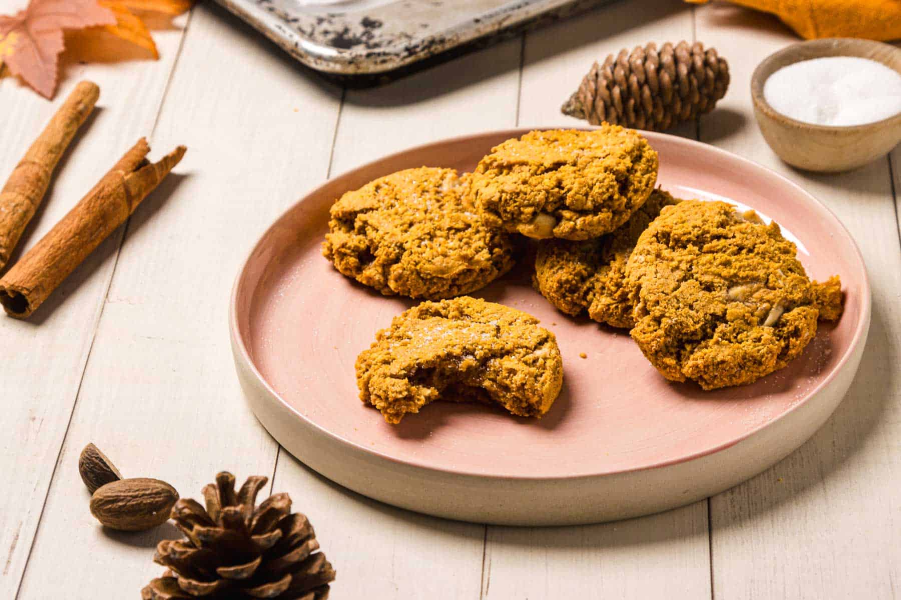 A plate of Pumpkin and White Chocolate Chip Cookies, one with a bite, is surrounded by pinecones, cinnamon sticks, and a bowl of sugar.