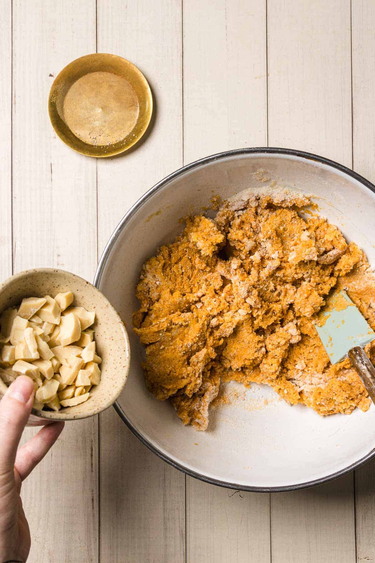 A hand holds a bowl of white chocolate chunks above pumpkin and white chocolate chip cookie dough in a mixing bowl with a spatula.