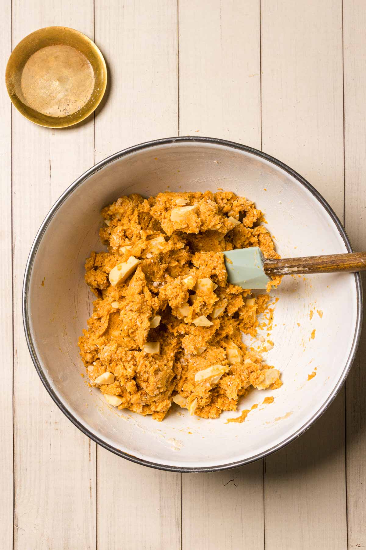 A bowl of cookie dough for Pumpkin and White Chocolate Chip Cookies, with white chocolate chunks and a spatula on a light wooden surface.