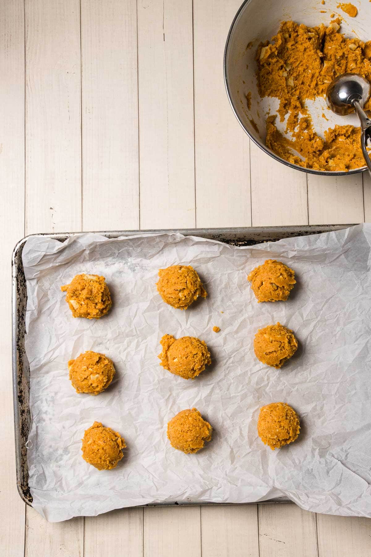 Nine scoops of Pumpkin and White Chocolate Chip Cookie dough on a parchment-lined baking tray; bowl with dough and scoop nearby.