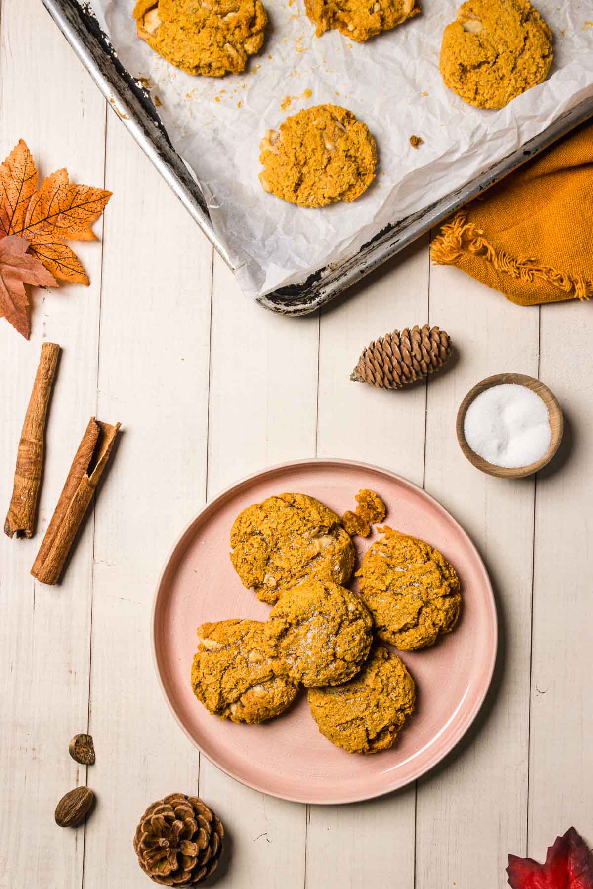 Pumpkin and White Chocolate Chip Cookies rest on a pink plate and baking tray, surrounded by autumn decor and cinnamon sticks.