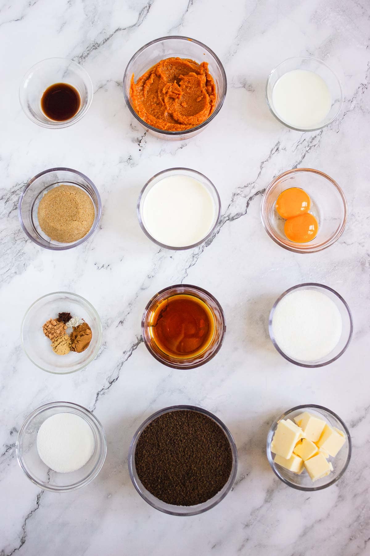 Twelve bowls with ingredients for Pumpkin Br&ucirc;l&eacute;e Tarts&mdash;pumpkin, sugar, eggs, butter, spices, milk, cream, and vanilla&mdash;arranged on a marble surface.