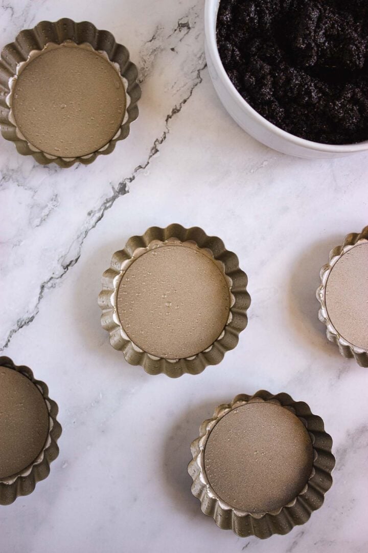 Five empty tart pans and a bowl of dark cookie crust mixture sit on a marble countertop, ready to be transformed into delicious Pumpkin Br&ucirc;l&eacute;e Tarts.