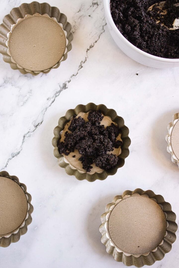 Five tart pans, including one chocolate-filled with crumb topping and others with plain filling, showcase delicious Pumpkin Br&ucirc;l&eacute;e Tarts on a marble surface.