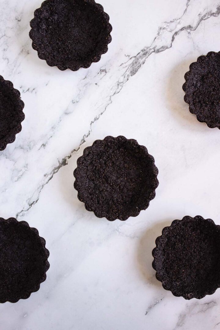 Six empty chocolate tart shells on a white marble surface, arranged in a scattered pattern&mdash;perfect bases for making delicious Pumpkin Br&ucirc;l&eacute;e Tarts.