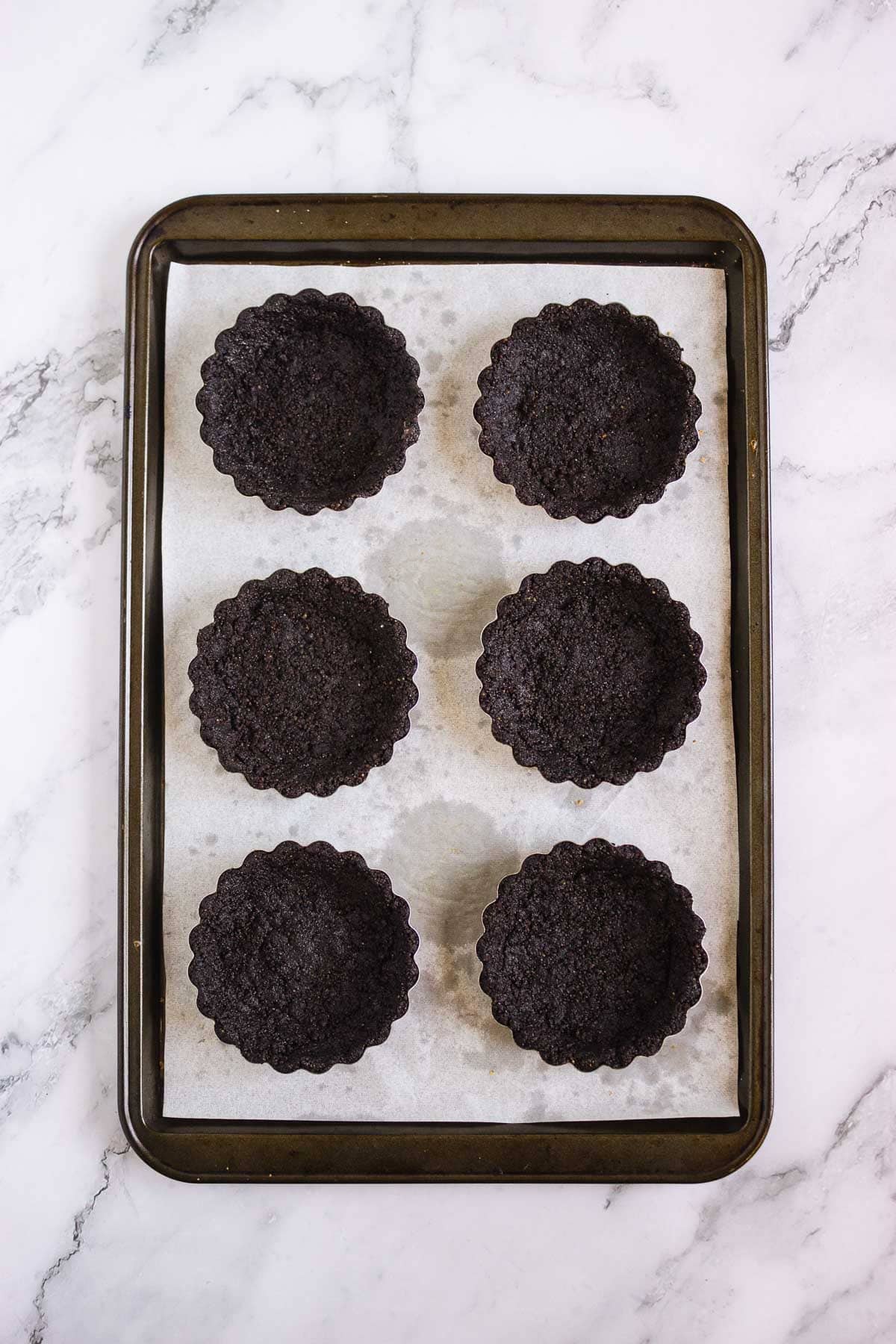 Six empty chocolate tart shells on a parchment-lined baking tray, viewed from above&mdash;perfect for creating delicious Pumpkin Br&ucirc;l&eacute;e Tarts.