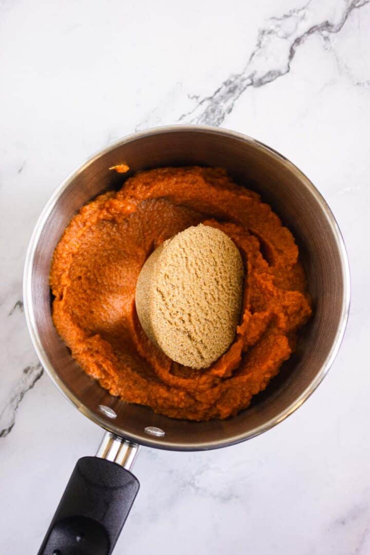A saucepan with pumpkin puree and a scoop of brown sugar on a white marble surface, ready to create delicious Pumpkin Br&ucirc;l&eacute;e Tarts.