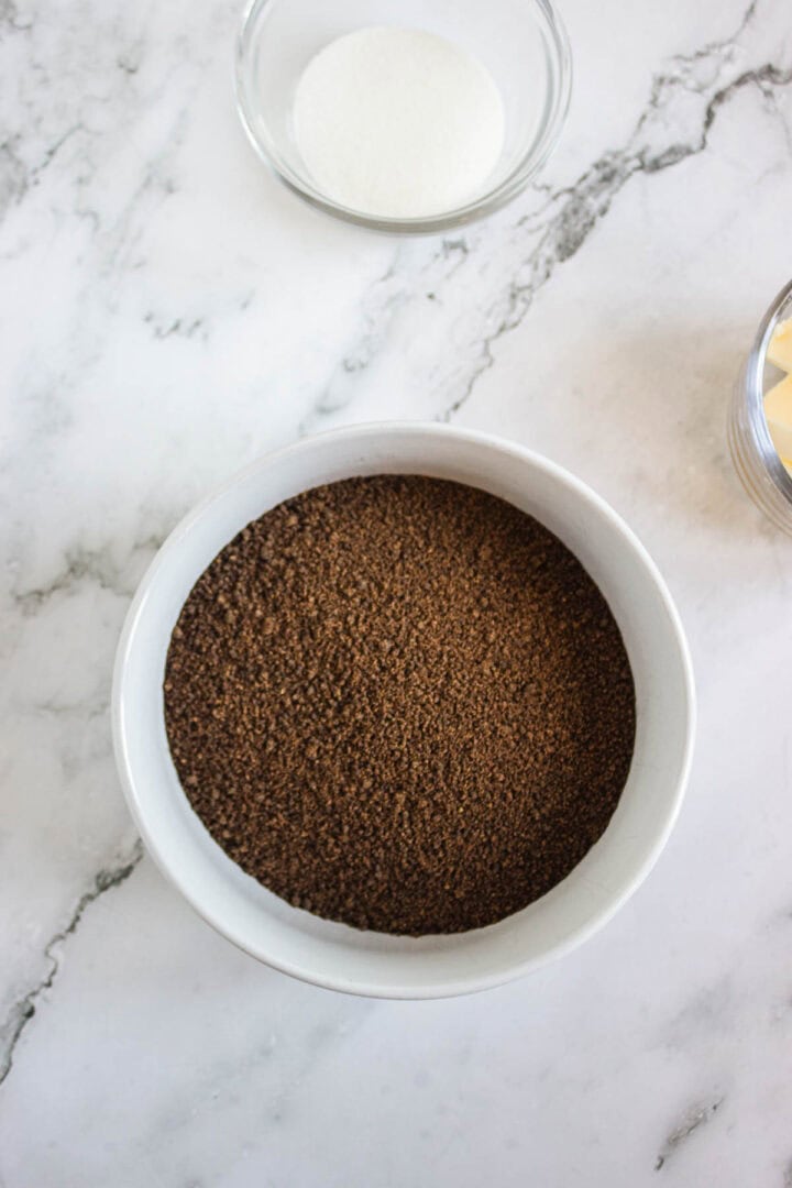 Bowl of crushed chocolate cookies on a marble surface, with sugar and butter in small bowls nearby&mdash;perfect for making Pumpkin Br&ucirc;l&eacute;e Tarts.