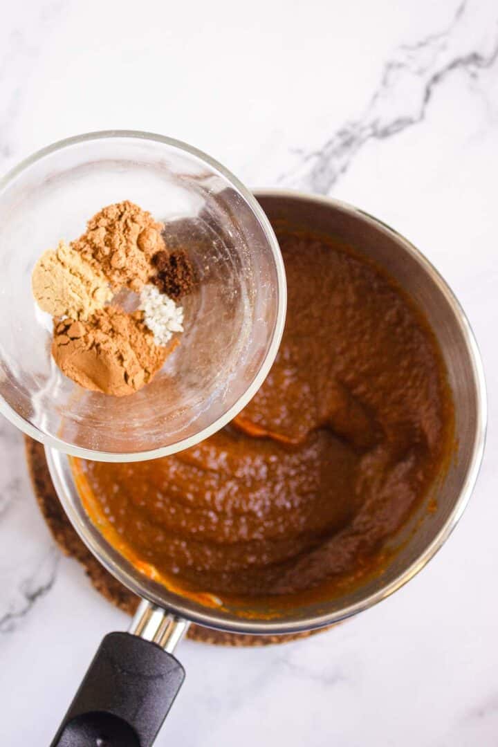 A bowl of spices is being held above a saucepan filled with pumpkin puree on a marble countertop, ready to create the filling for delicious Pumpkin Br&ucirc;l&eacute;e Tarts.
