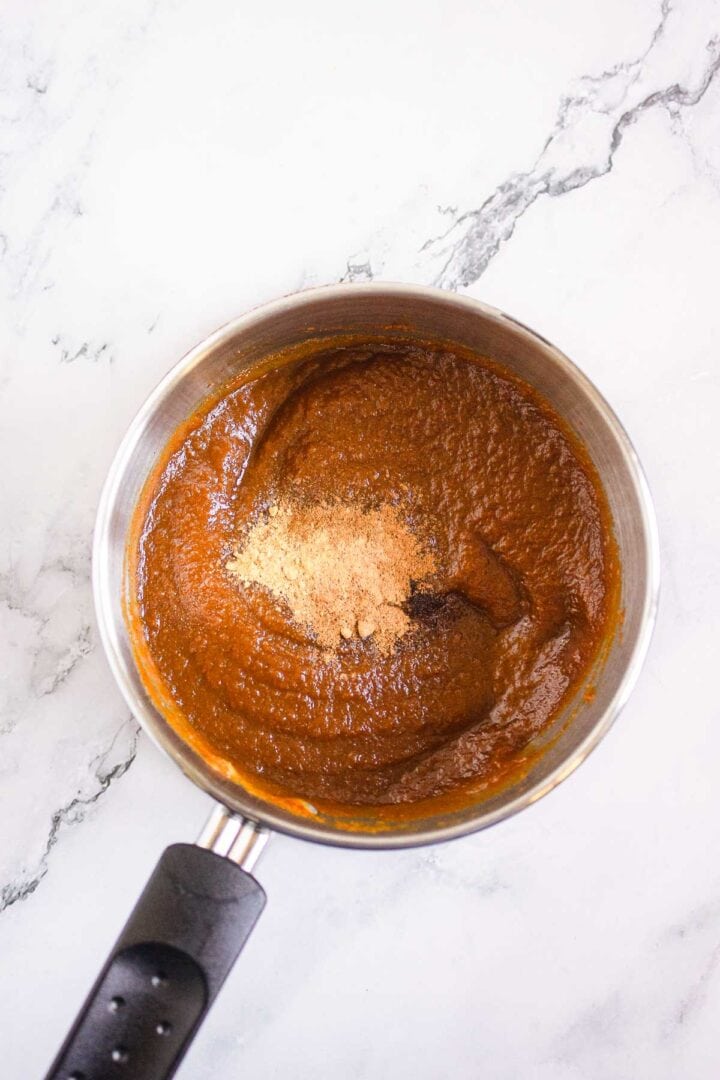 A saucepan with thick brown sauce and spices on top, reminiscent of the caramelized topping on Pumpkin Br&ucirc;l&eacute;e Tarts, placed on a white marble surface.