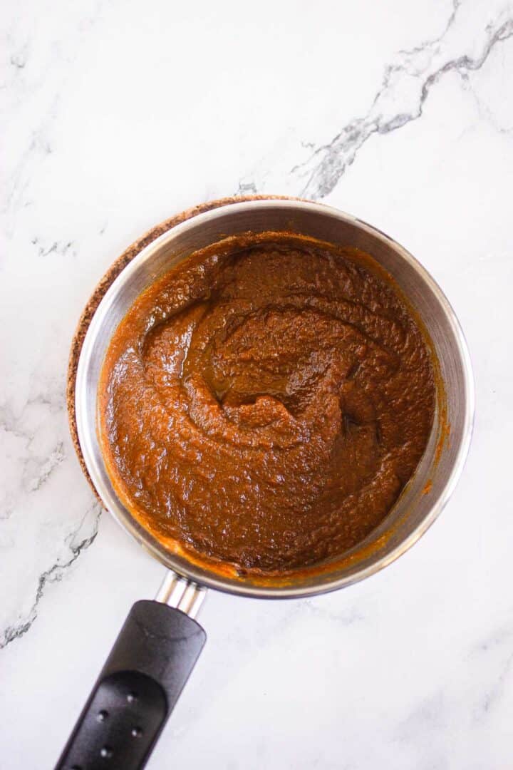 A saucepan filled with thick, dark brown sauce sits on a white marble surface, ready to drizzle over Pumpkin Br&ucirc;l&eacute;e Tarts.