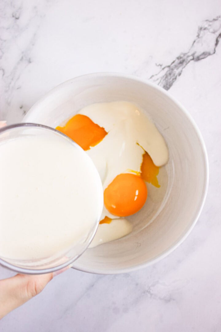 A bowl with egg yolks and cream, perfect for preparing Pumpkin Br&ucirc;l&eacute;e Tarts, with more cream being poured in from another bowl on a marble counter.