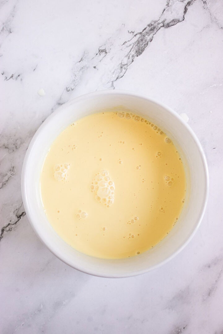 A white bowl filled with a smooth, light yellow liquid mixture on a marble surface, perfect for creating delicious Pumpkin Br&ucirc;l&eacute;e Tarts.