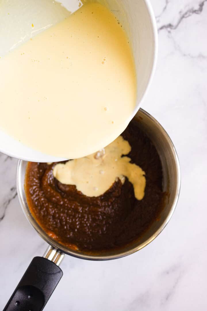 Custard mixture being poured from a bowl into a saucepan of dark puree on a marble countertop, the perfect start for luscious Pumpkin Br&ucirc;l&eacute;e Tarts.