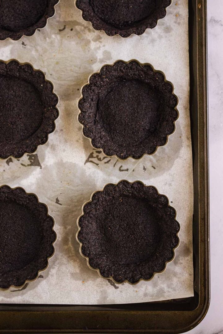 Six empty tart crusts made with a dark chocolate cookie base on a parchment-lined baking sheet, ready to be filled for Pumpkin Br&ucirc;l&eacute;e Tarts.