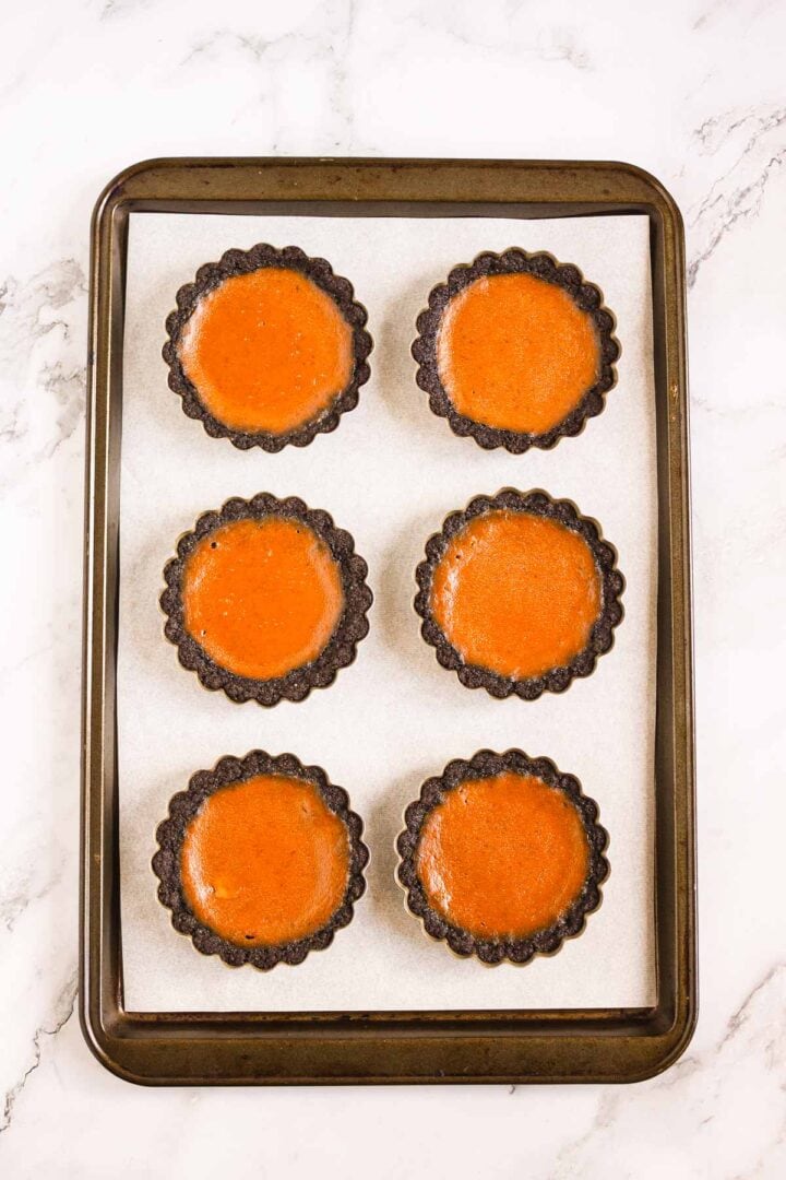 Six Pumpkin Br&ucirc;l&eacute;e Tarts with dark crusts rest on a parchment-lined baking sheet, displayed on a marble surface.