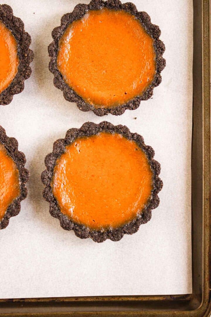 Three Pumpkin Br&ucirc;l&eacute;e Tarts with dark chocolate crusts rest on a parchment-lined baking sheet, their caramelized tops adding an elegant touch.