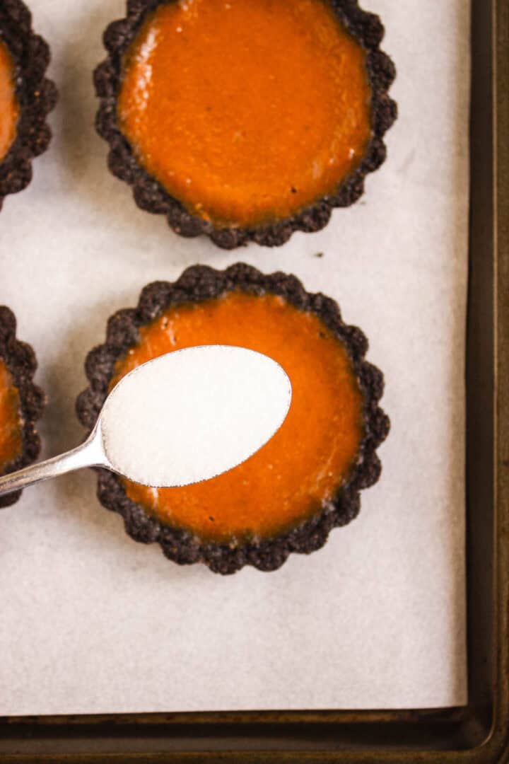 A spoon of sugar held above Pumpkin Br&ucirc;l&eacute;e Tarts with an orange filling and a chocolate crust on a baking tray.